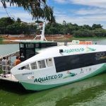 Barco "Bororé I" durante demonstração na Represa Billings. O barco está visto na diagonal dianteira sobre a água esverdeada. Ao lado há uma plataforma de embarque flutuante. Há algumas pessoas na parte dianteira do barco. Ao fundo pode ser vista uma margem.